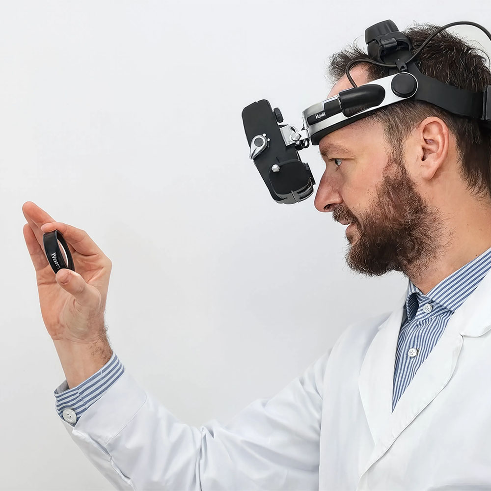 Ecco l'alt text per questa immagine: "Medico oculista con visore binoculare Keeler indossato sulla testa esamina un piccolo dispositivo che tiene in mano, sfondo neutro