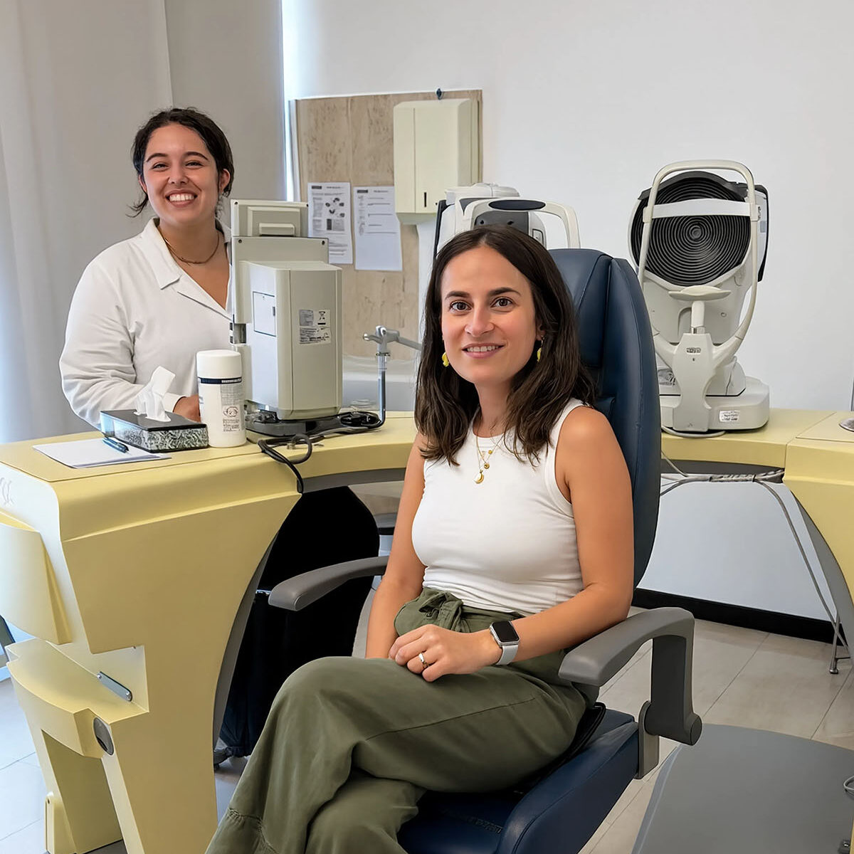 Paziente sorridente seduta sulla poltrona per la visita oculistica, con oftalmologa sullo sfondo circondata da strumentazione diagnostica