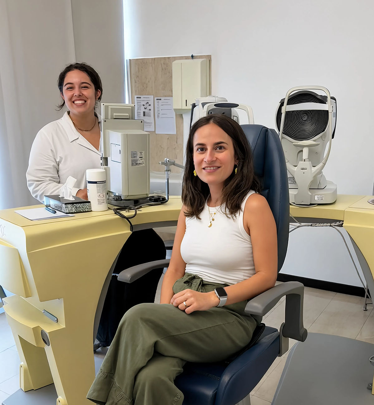Paziente sorridente seduta sulla poltrona per la visita oculistica, con oftalmologa sullo sfondo circondata da strumentazione diagnostica