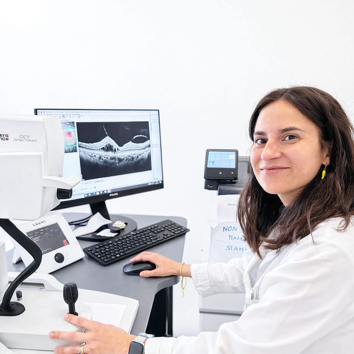 Oculista sorridente seduta alla strumentazione OCT durante l'esame della retina, con scansione visibile a monitor