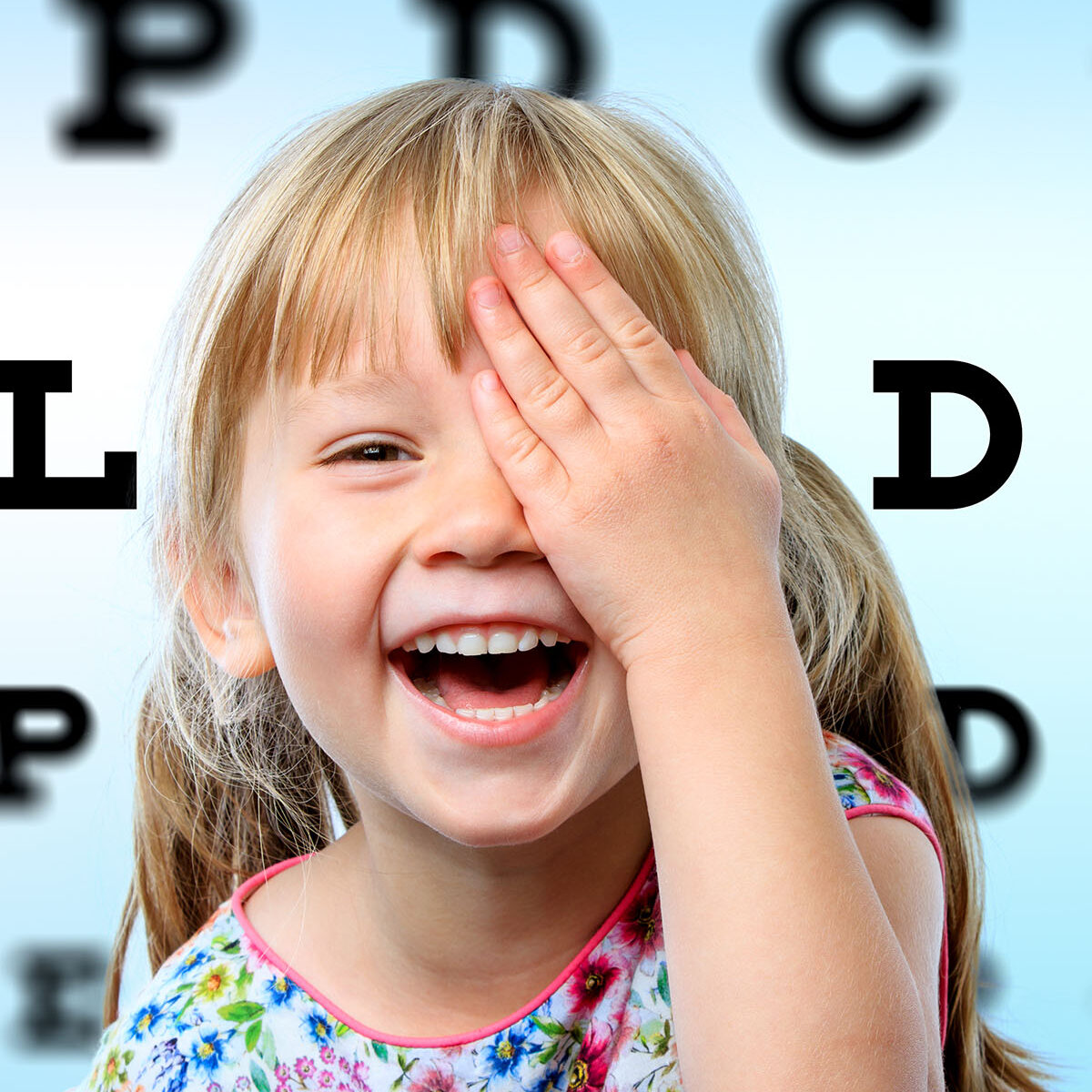 Bambina sorridente si copre un occhio con la mano durante il test della vista, con tabella optometrica sfocata sullo sfondo